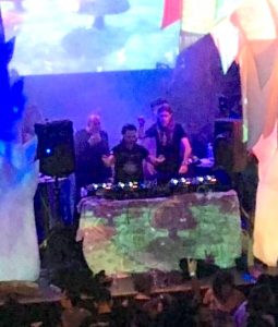 Bathed in colored lighting, an electronic music band performs on a stage while a screen hanging above them displays swirling images. In the foreground of the photo can be seen audience members standing shoulder to shoulder in front of the stage.