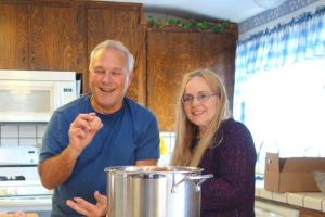 A smiling man triumphantly holds up a prepared meatball before cooking it. Alongside him, a blonde woman also smiles for the camera. Both are posing behind a large pot of homemade tomato sauce. 