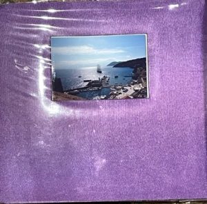 A homemade scrapbook is covered in plush velvety purple fabric. In the center of the book cover is mounted a scenic photograph of a port in Italy. Boats, water, blue sky, and fluffy clouds are all visible in the background of the photograph.