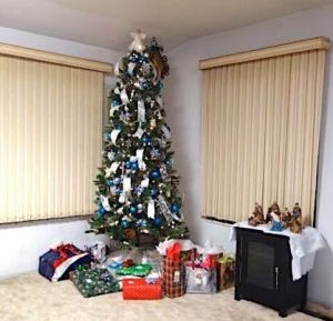 A fully decorated Christmas tree, festooned with ribbons and ornaments with an angel on top, is on display in the corner of a living room. Piles of wrapped presents are stacked below the tree. 