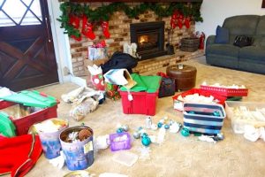 A selection of Christmas ornaments are spread across a beige carpet in a living room. Some ornaments are unwrapped and others are still packed into storage containers. In the background, a fire is burning in a brick fireplace that is decorated with an evergreen garland and red Christmas stockings.