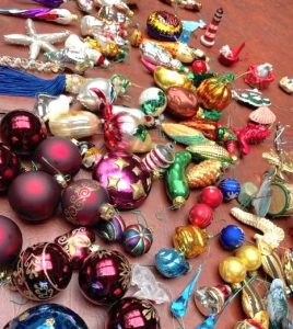 A selection of vintage glass Christmas ornaments is spread across a table. There are classic round ornaments visible alongside ones shaped like birds, sea life, and vegetables. 