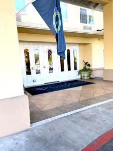 The front entrance of a Carlton senior living facility shows white doors with glass windows, bright yellow exterior wall paint, and tiled pavers in front of the doors.