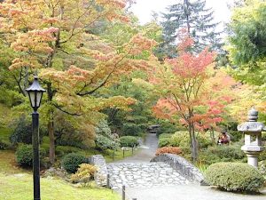 In the foreground is a stonework walking bridge, sculpture, and an iron lamp post, while the background shows a paved walking path and beautiful shrubbery and trees in shades of green, yellow, orange, and red.