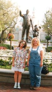 Two women pose in front of the bronze statue of Walt Disney and Mickey Mouse on the grounds of Disneyland. Cinderella's castle can be seen behind them in the distance.