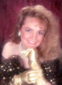 A stylized 1980s studio glamour portrait shows a smiling blond woman wearing gold metallic formal gloves and a black, off-the-shoulder dress patterned with metallic gold polka dots.