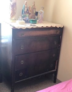 A dark wood dresser sits in a corner of a bedroom near a window. On top is a square lace doily, on which is placed a couple of small trinket boxes and a collection of ceramic statues of religious figures.
