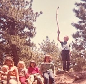 During a camping trip in the woods, five children pose for a photo by sitting on the trunk of a fallen tree, while another boy holds up a big stick.