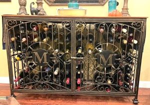A collection of wine bottles is displayed and stored inside a type of decorative, locking rack known as a "wine jail." It features elaborate filigree metal doors and monogrammed medallions on the front.