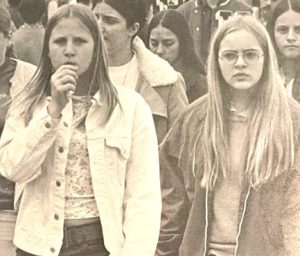 A vintage sepia-toned photo shows two teenage girls. The girl on the left has straight, dark blonde hair and is wearing a light-colored jacket with buttons. The girl on the right has longer, lighter blonde straight hair, and is wearing glasses.