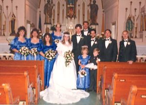 A slightly blurry vintage photograph shows a 1980s church wedding party; the bride wears a long-sleeved white gown with train and a headband with veil, while the groom wears a black tuxedo. The four groomsmen and ring bearer also wear black tuxes, while the four bridesmaids and a flower girl wear royal blue satin gowns. All of the women hold large flower bouquets.
