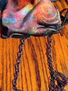 A cloth face mask made from black fabric is printed with a rainbow pattern that resembles astral photographs. Attached is a rainbow chainmail chain.