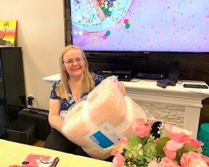 A blonde woman in glasses and a blue-patterned shirt is seated at a table behind a large bouquet of pink roses. In her hands is a birthday gift she was just given: a large, fuzzy, pale pink blanket in clear packaging.