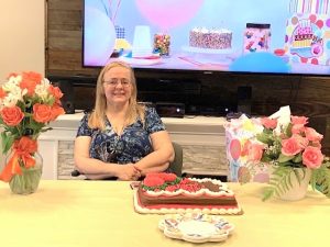 A blonde woman in glasses and a blue-patterned shirt is seated at a table behind a large birthday cake decorated with pink icing flowers. On either side of the cake are two bouquets of roses in vases