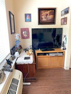 A cozy corner of a shared room in a memory care facility shows a large window on one side, plus nice plank flooring, butter-colored painted walls decorated with framed photographs and artwork, and a flatscreen TV on a cabinet.