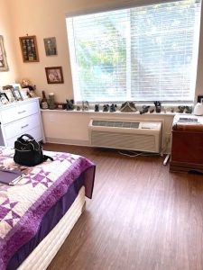 A cozy dorm-style room features a bright picture window covered in blinds, with a wide ledge in front on which statues, candles, and other personal decorative items are arranged. The room also features plank flooring, painted walls, and a dresser on the left with lots of framed photos.
