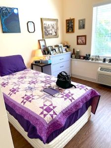 A cozy dorm-style room has a bright window, plank flooring, butter-colored painted walls decorated with a few paintings, and a dresser with lots of framed photos. On the bed is a beautiful ivory quilt featuring a bright purple quilted geometric pattern.
