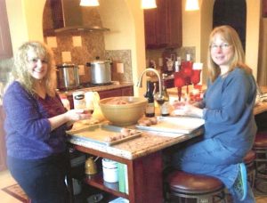 Two blonde women, in the process of making meatballs at a large kitchen island, smile at the camera.
