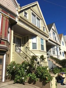 A woman sits on the steps of an olive green Victorian house in San Francisco. There is a hedge of green bushes in the foreground.