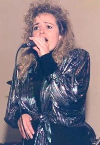 A curly-haired blonde women wearing a metallic blouse sings into a microphone.