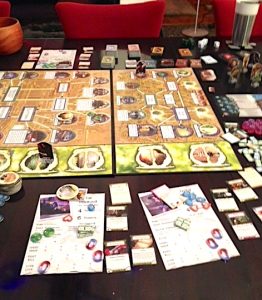 The Arkham Horror board game is shown elaborately set up on a table. Next to the board on the tabletop are a variety of player pieces, game tokens and stacks of game cards.