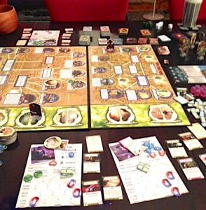 The Arkham Horror board game is shown elaborately set up on a table. Next to the board on the tabletop are a variety of player pieces, game tokens and stacks of game cards. 