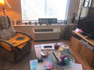 A cozy carpeted living room shows a wide picture window with an upholstered chair, floor lamp, wooden coffee table, wooden TV cabinet, and large flatscreen TV.