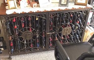 Peggy's fully stocked wine jail (a decorative cabinet wine rack with locking filigree metal doors) is shown in its new home in her assisted living apartment's living room. Resting on top are framed family photos.