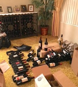 A couple of dozen bottles of wine are being packed up next to a stocked wine jail (a decorative rack that locks). The sorted bottles are arranged in piles on a mustard yellow living room carpet, next to special packing boxes.