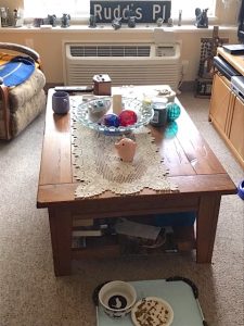 A wooden coffee table in the center of a cozy living room. On top are a cream lace runner, a glass bowl of decorative glass spheres, a mug, and a ceramic pig figurine.