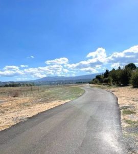 Against a bright blue sky dotted with low, fluffy white clouds, a wide, curving paved trail road stretches out to the horizon. Grass is visible in the foreground on either side of the road, and lots of trees visible in the background. 