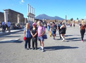 Against a bright blue sky, three tourists pose in front of the historic site of Pompeii. Mount Vesuvius is in the background at the horizon, while many other visitors can be seen walking around.
