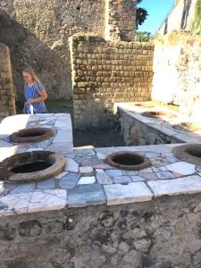 A blonde woman visits the site of an ancient "fast food" stand in Pompeii, essentially a stone-and-tile countertop with embedded jars.