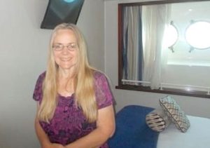A smiling blonde woman in a purple shirt is sitting on the bed in a modest cruise ship stateroom. Bright sun shines through two portal windows behind her.