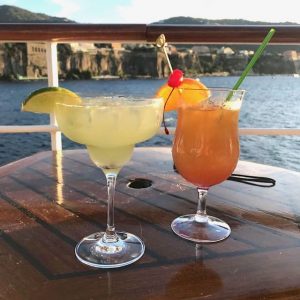 Two fruity cocktails sit on a table; in the background is the railing of a cruise ship, water, and a beautiful skyline of a port in Italy.