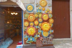 A view of a shop doorway in Sicily. To the right of the doorway is a display of bright yellow, orange, and turquoise wall decorations, most of which are sun faces.