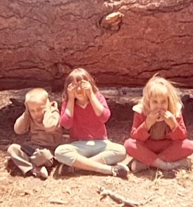 Vintage photo of a boy and two girls seated in front of a fallen tree at a campground in the woods. The boy is covering his ears with his hands, the girl in the center is covering her eyes, and the girl on the right is covering her mouth. 
