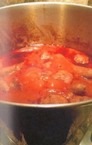 A close-up photo of a huge silver lobster pot filled with bubbling red spaghetti sauce, meatballs, and sausages.