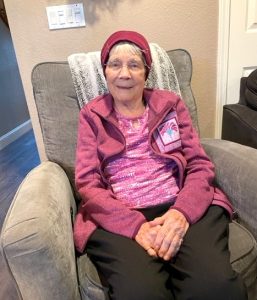 An elderly, smiling woman is shown seated on a plush grey upholstered armchair. She is wearing a pink top, a comfortable pink lounge jacket, and a pink hat.