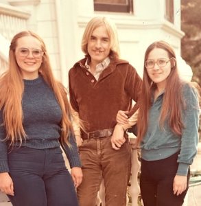 A vintage snapshot depicting a young woman with long strawberry blonde hair and glasses, a young man with collar-length blonde hair, and a young woman with long brown hair and glasses.