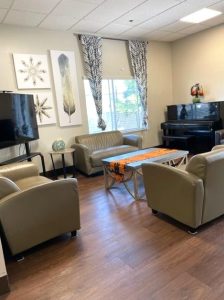 A cozy living room has a bright window, plank flooring, beige walls decorated with a few paintings, an upright piano, a flat-screen TV, a leather loveseat and wide arm chairs, and a coffee table.