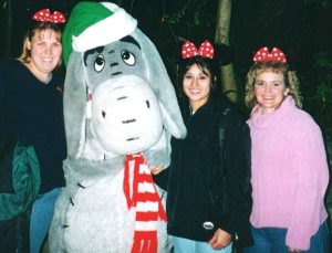 Three women pose with the costumed character of Eeyore at Disneyland. Each woman wears mouse ears decorated with a red, polka-dotted bow. Eeyore wears a green and white "santa" hat and a red and white striped winter scarf. 