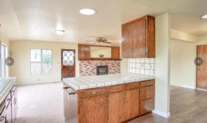 A view of a carpeted family room while standing in the adjacent kitchen. Windows, a glass-topped exterior door, and a bricked fireplace stand on the far wall.