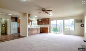 This view of a freshly carpeted living room shows a kitchen in the background with new plank flooring, as well as a sliding glass door through which a backyard can be seen. The left of the photo shows a hallway to the rest of the house.