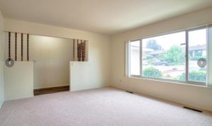 This view of a living room shows a large front picture window to the right, through which greenery and a neighbor's house can be seen, and brand new beige carpeting. In the back left of the photo is the house's entrance hall with new plank flooring, and the front door.