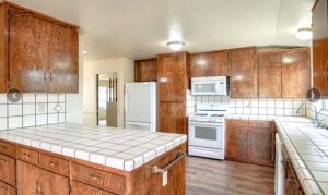 A kitchen view shows dated dark wood cabinets, white-tiled countertops with dark grout, and white appliances. The plank flooring is brand new.