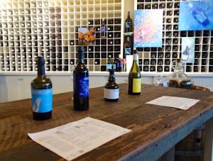 In the foreground is a handcrafted wooden countertop. Displayed on top are several open bottles of wine, ready for tasting. In the background is a unique-looking wine rack wall, where a grid of narrow shelves holds many bottles.
