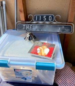 A clear plastic storage bin with blue handles holds many folders of documents. On top of the box is a ring of keys and a CD; behind the box is a decorative metal address sign.