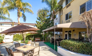 A courtyard of a large building shows tables with umbrellas, a covered patio, and palm trees.