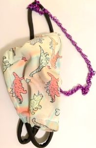 A cloth face mask made from beige fabric that is printed with colorful drawings of various dinosaurs. It sports black ear loops and a purple chainmail chain.
